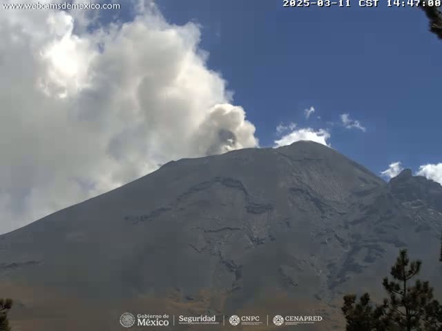 A large, dark-grey volcano emits a plume of light-grey smoke under a mostly clear, bright blue sky.