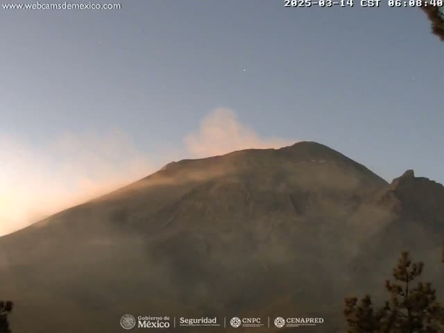 A large, dark-colored mountain is partially obscured by a light-brown plume of ash or dust under a clear, pale-blue sky.