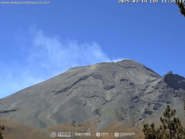 A large, gray volcanic mountain emits a thin plume of smoke under a clear blue sky.