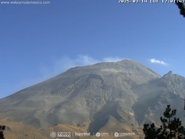 A large, gray volcanic mountain is seen on a clear, sunny day with a small amount of smoke or ash emanating from its peak.