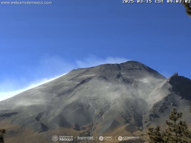A large, gray volcanic mountain is partially obscured by a light dust cloud under a clear blue sky.