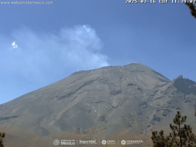 A large, dark-gray volcano with a plume of light-gray smoke rising from its peak is visible under a clear blue sky.