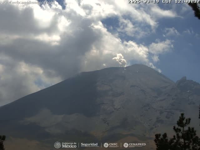 A large, dark-grey mountain with a plume of smoke or ash erupting from its peak is visible under a partly cloudy sky.