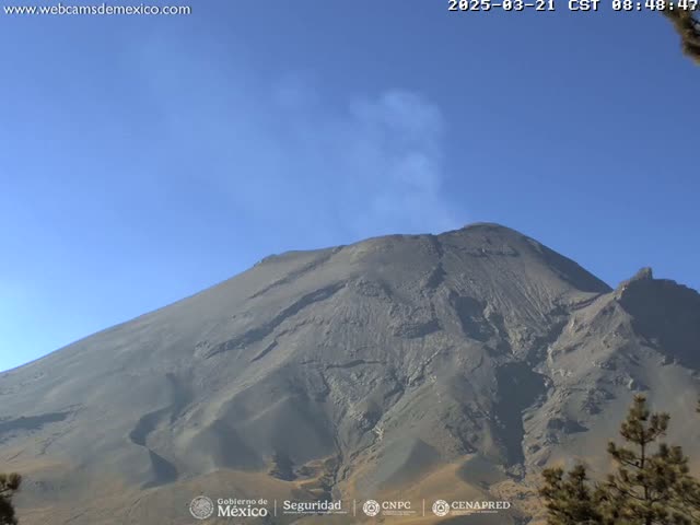 A large, dark-gray volcano with a small plume of smoke emanating from its peak is visible under a clear blue sky.