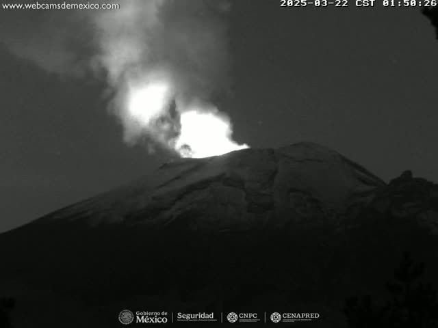 A nighttime, black and white image shows a volcano erupting with bright light and smoke visible against a dark sky.
