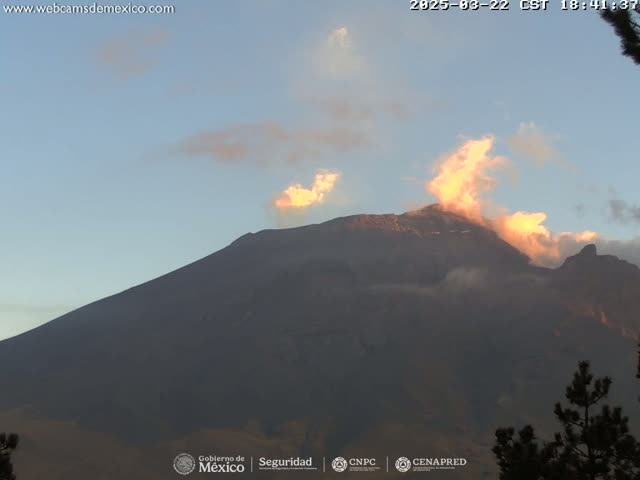A large, dark mountain is partially shrouded in clouds, with a plume of light orange smoke rising from its peak under a mostly clear, pale blue sky.