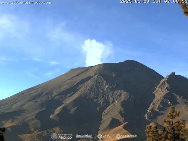 A dark-colored volcano emits a plume of white smoke under a clear blue sky.