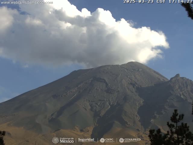 A large, dark-grey volcano is partially obscured by a large white cloud under a mostly clear blue sky.