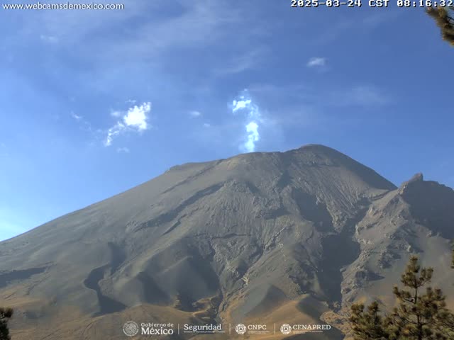 A large, dark gray volcano emits a plume of light-gray smoke under a mostly clear, blue sky.