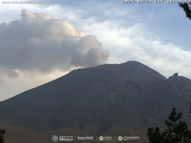 A large, dark mountain with a plume of smoke or ash rising from its peak is visible under a partly cloudy sky.