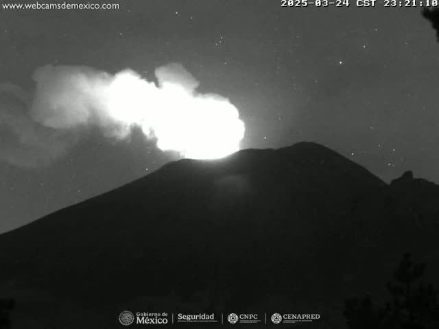 A nighttime, low-light image shows a volcano erupting with a bright plume of smoke and ash against a starry sky.