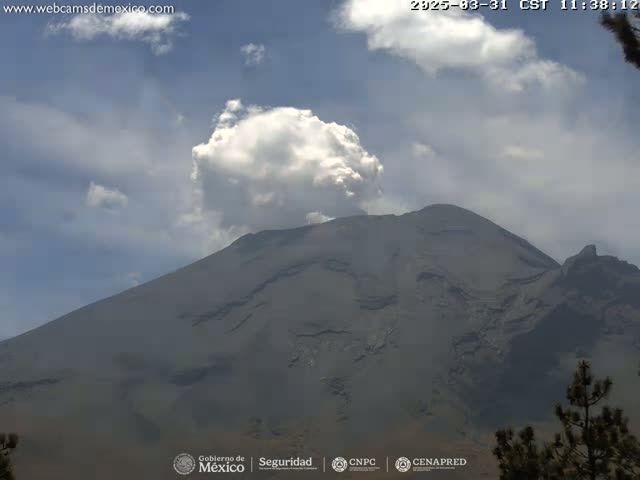 A large, dark-grey volcano with a plume of white smoke erupting from its summit is visible under a partly cloudy sky.