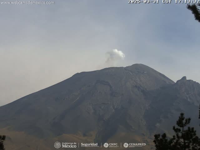 A large, dark-gray volcano with a small plume of white smoke erupting from its peak is visible under a hazy sky.