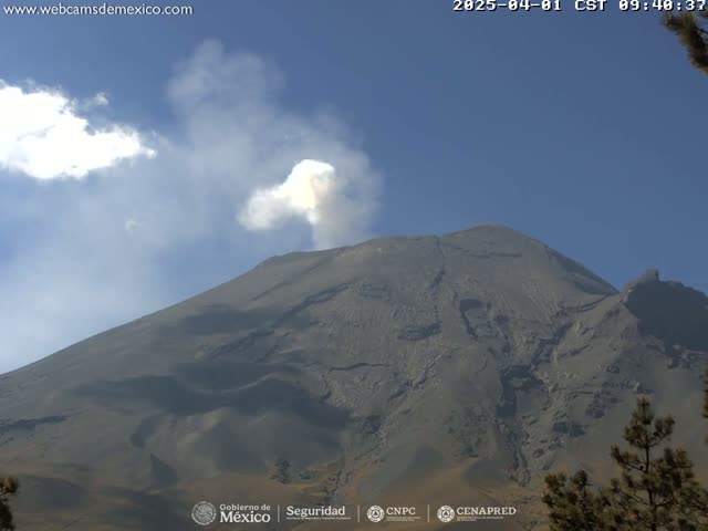 A large, dark-grey volcano with a plume of light-grey smoke erupting from its peak under a mostly clear blue sky.
