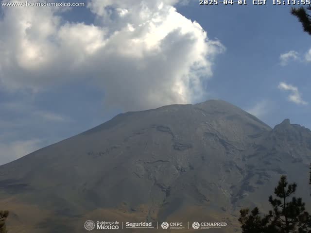 A large, dark-gray volcano with a plume of smoke or steam rising from its peak is visible under a partly cloudy sky.