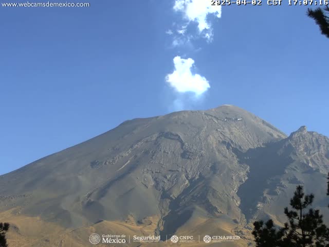 A large, gray volcano with a small plume of white smoke erupting from its peak is visible under a clear blue sky.