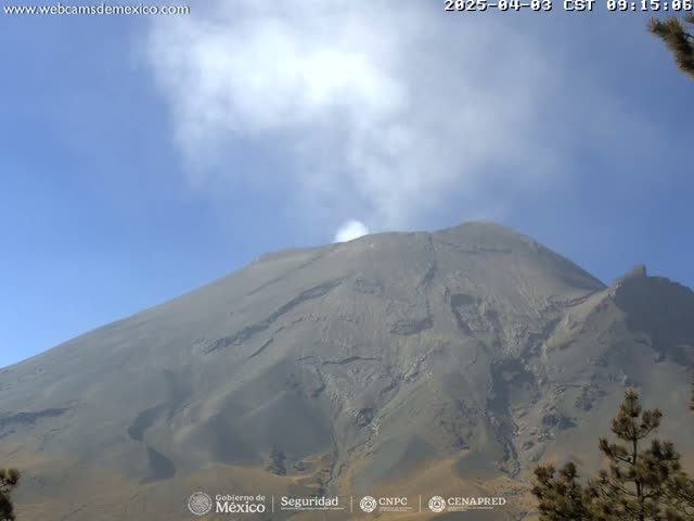 A large, gray volcano with a plume of smoke emanating from its peak is visible under a clear, blue sky.