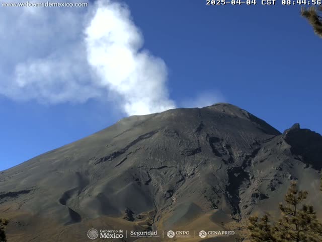 A large, dark-colored volcano emits a plume of white smoke under a clear blue sky.
