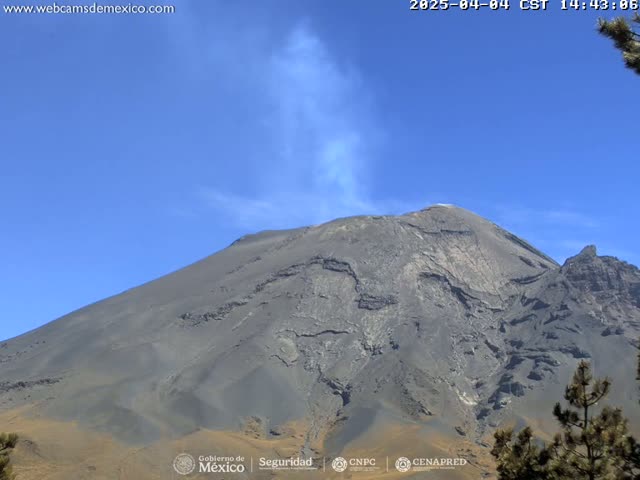 A large, gray volcano emits a plume of light-colored smoke under a clear blue sky.