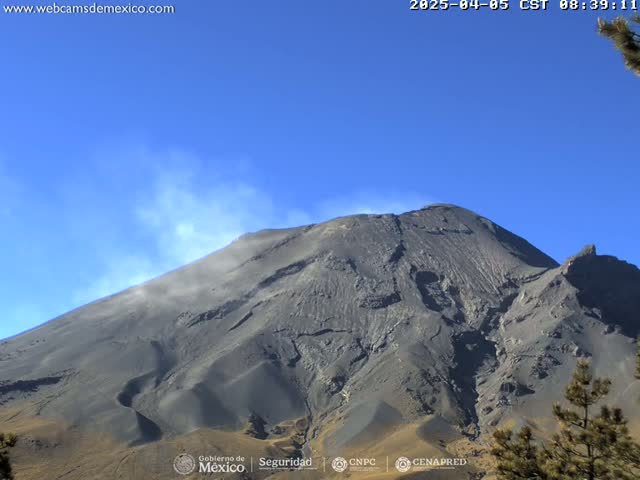 A large, dark-grey volcanic mountain with a plume of light-grey ash rising from its side under a clear blue sky.
