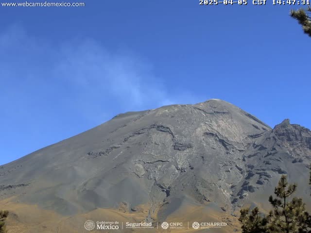 A large, dark gray volcano with a plume of light gray smoke rising from its summit is visible under a clear blue sky.