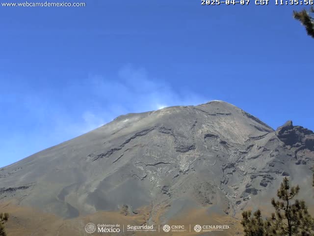 A large, gray volcano with a small plume of smoke erupting from its summit is visible under a clear blue sky.