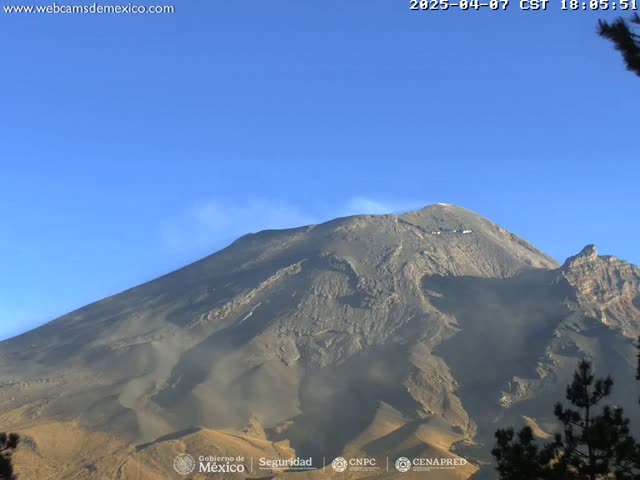 A large, dark-colored volcano with a plume of smoke or ash emitting from its peak is visible under a clear blue sky.