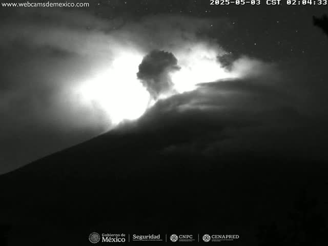 A nighttime, low-light image shows a volcano erupting, emitting bright light and dark smoke or ash into the night sky.