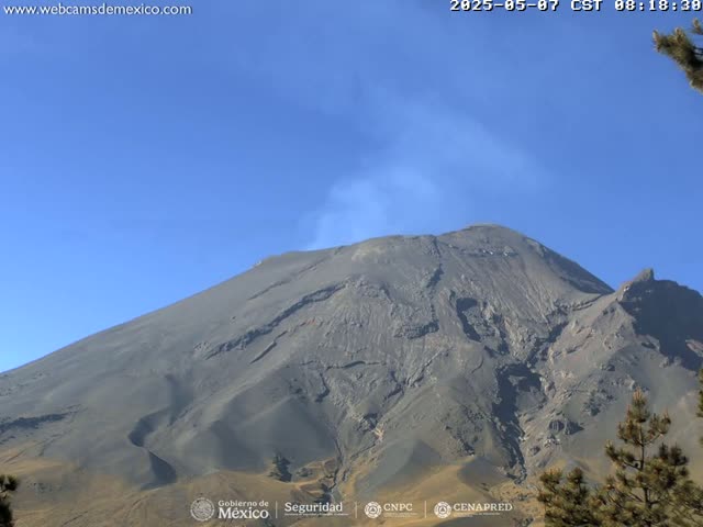 A large, dark gray volcano with a plume of smoke rising from its peak is visible under a clear blue sky.