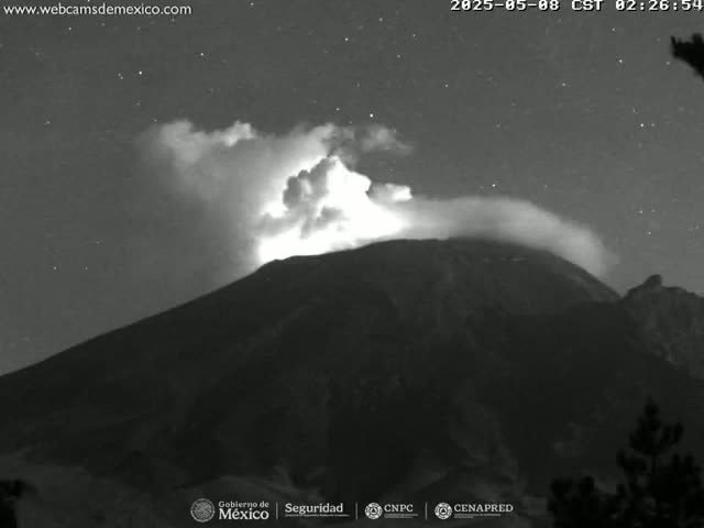 A nighttime, grayscale image shows a volcano emitting a plume of smoke or ash under a starry sky.