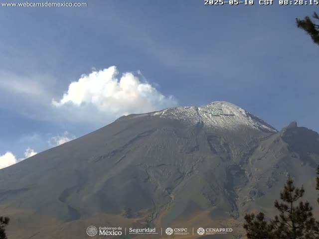 A large, snow-capped volcano is seen under a mostly clear, blue sky with a few puffy clouds.