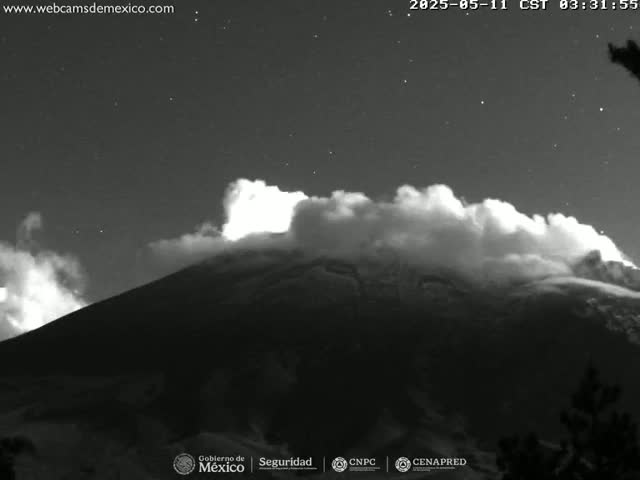 A nighttime, black and white image shows a volcano erupting, with a large plume of smoke or ash rising above it under a starry sky.