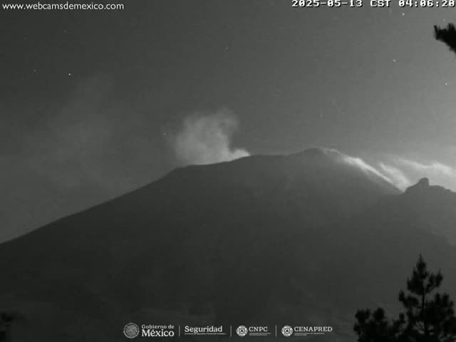 A dark, nighttime view shows a volcano emitting a plume of smoke or ash under a mostly clear sky.