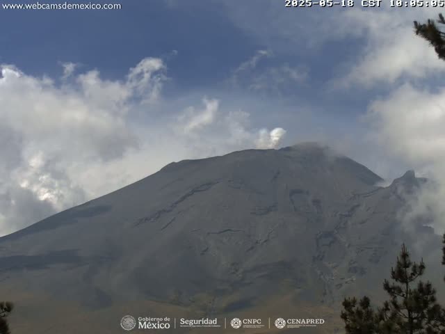 A large, gray volcano with a small plume of smoke erupting from its peak is partially obscured by partly cloudy skies.
