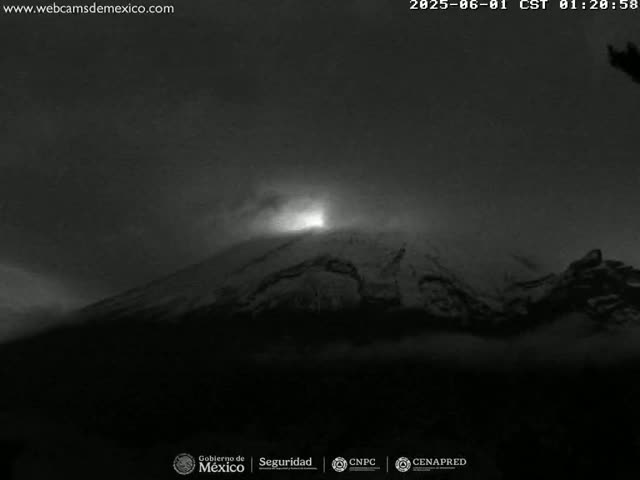 A nighttime, low-light image shows a snow-covered mountain with a bright light emanating from its peak under a cloudy sky.