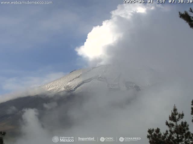 A snow-capped mountain is partially obscured by clouds and fog under a partly sunny sky.
