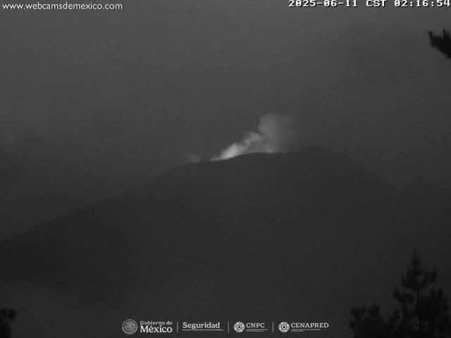 A nighttime, hazy image shows a volcano emitting a plume of smoke or ash.