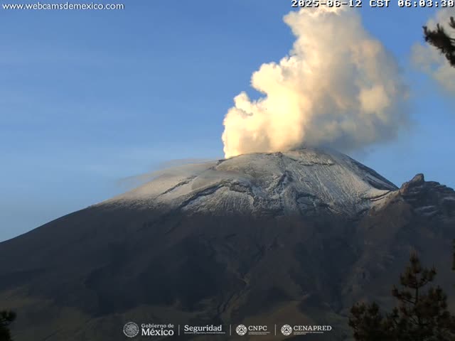 A snow-capped volcano erupts a large plume of smoke and ash under a clear blue sky.