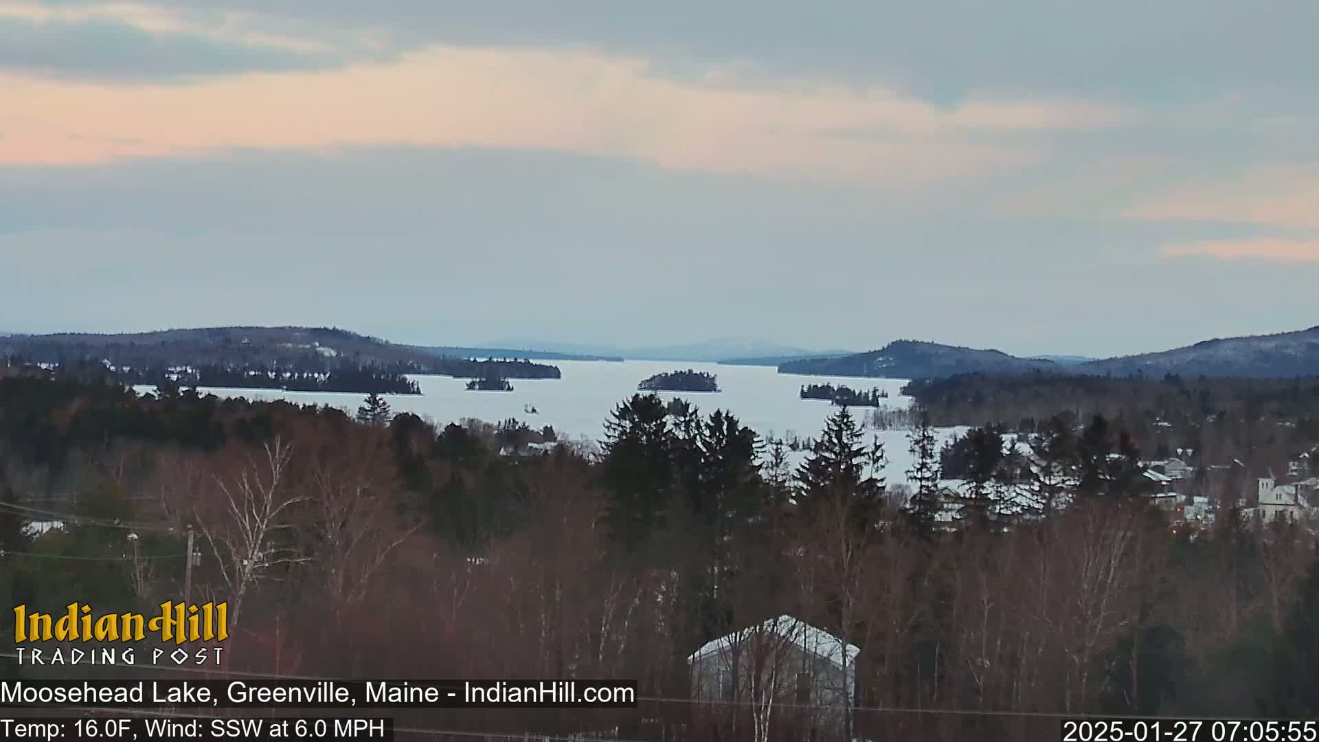 A snow-covered lake with several islands is seen from a wooded hillside under a mostly cloudy sky.