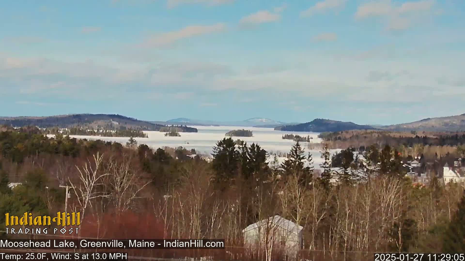 A snow-covered lake stretches to distant mountains under a partly sunny sky, with a village nestled in the foreground among leafless trees.