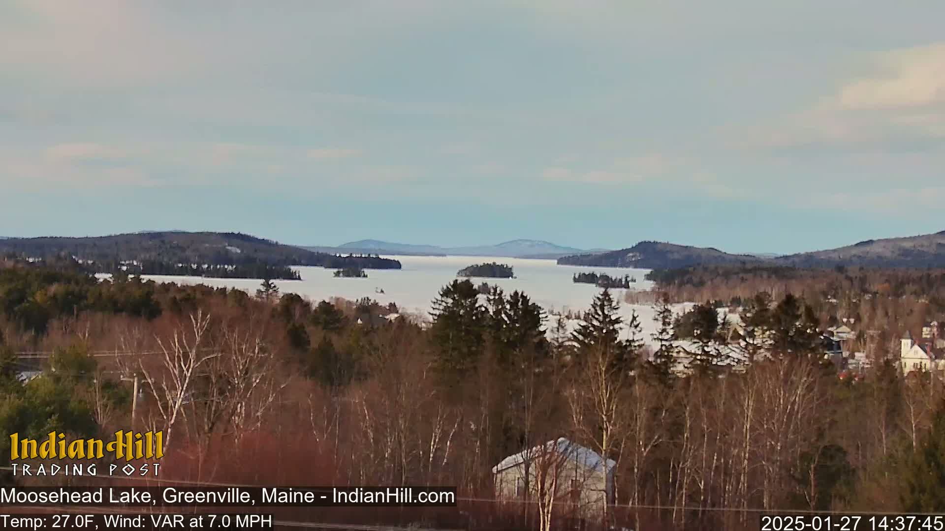 A snow-covered lake stretches between wooded hills under a mostly clear sky.