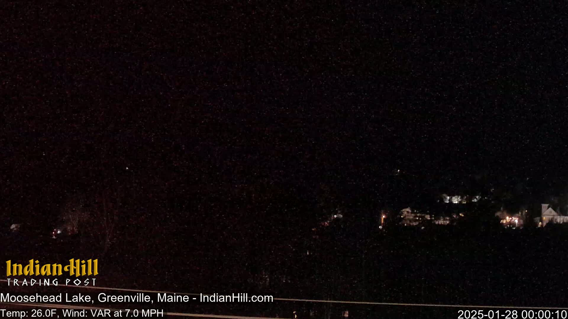 A nighttime view of a dark landscape with a few lights visible in the distance under a starlit sky, with a temperature of 26°F and a variable wind at 7.0 mph.