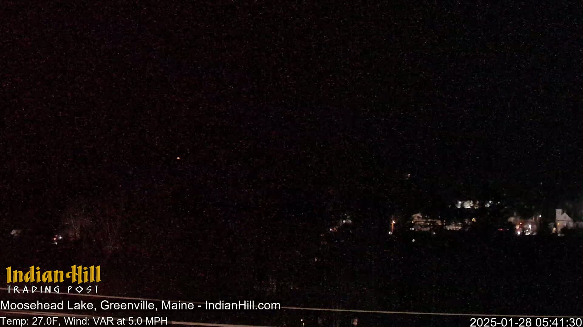A nighttime long shot shows a dark, snow-filled sky over a dimly lit town under a variable wind at 5.0 mph and a temperature of 27 degrees Fahrenheit.