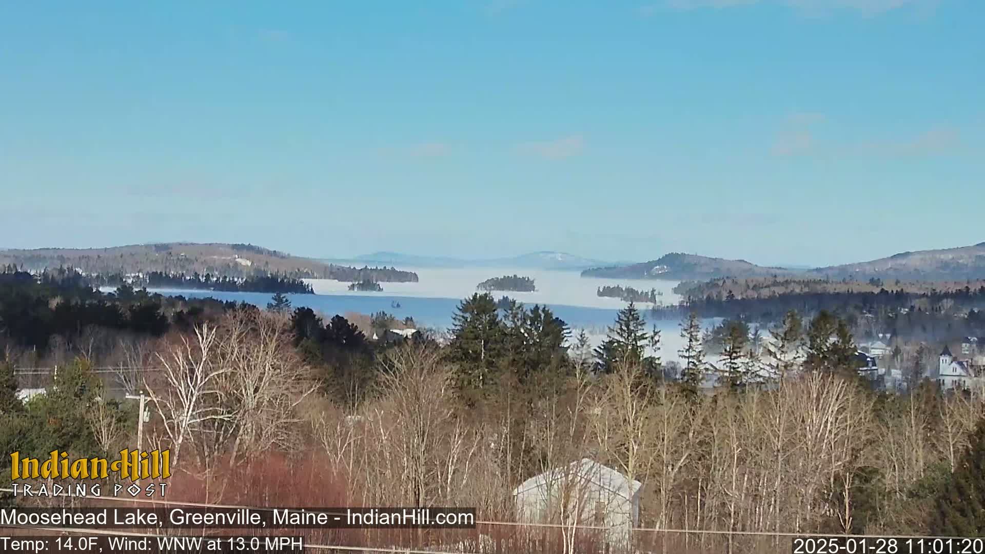 A large lake covered in fog is seen from a distance, surrounded by snow-covered hills and bare winter trees under a clear blue sky.