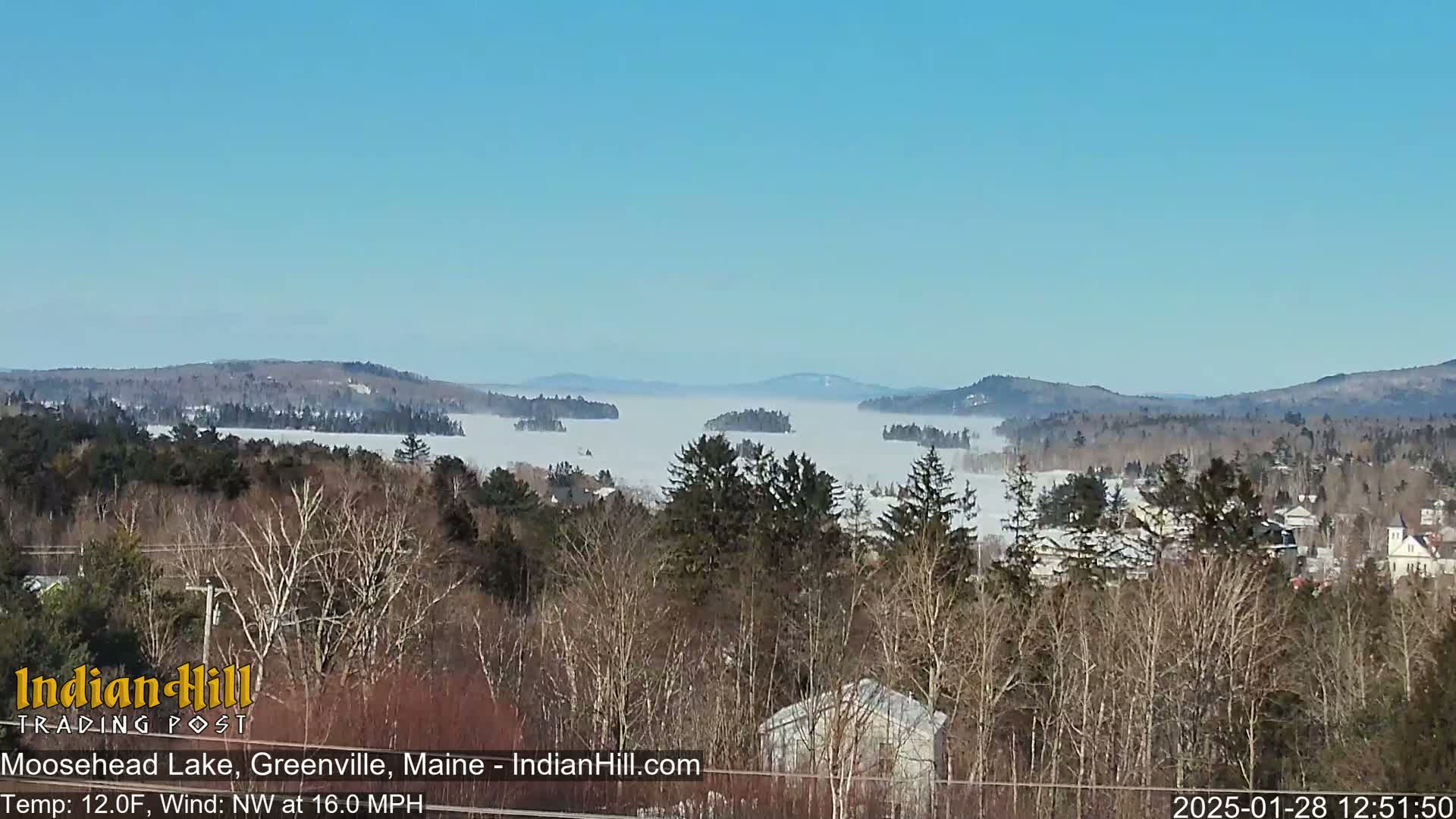 A mostly clear, cold day shows a view of a lake covered in fog, with snow-dusted trees and distant hills in the background.