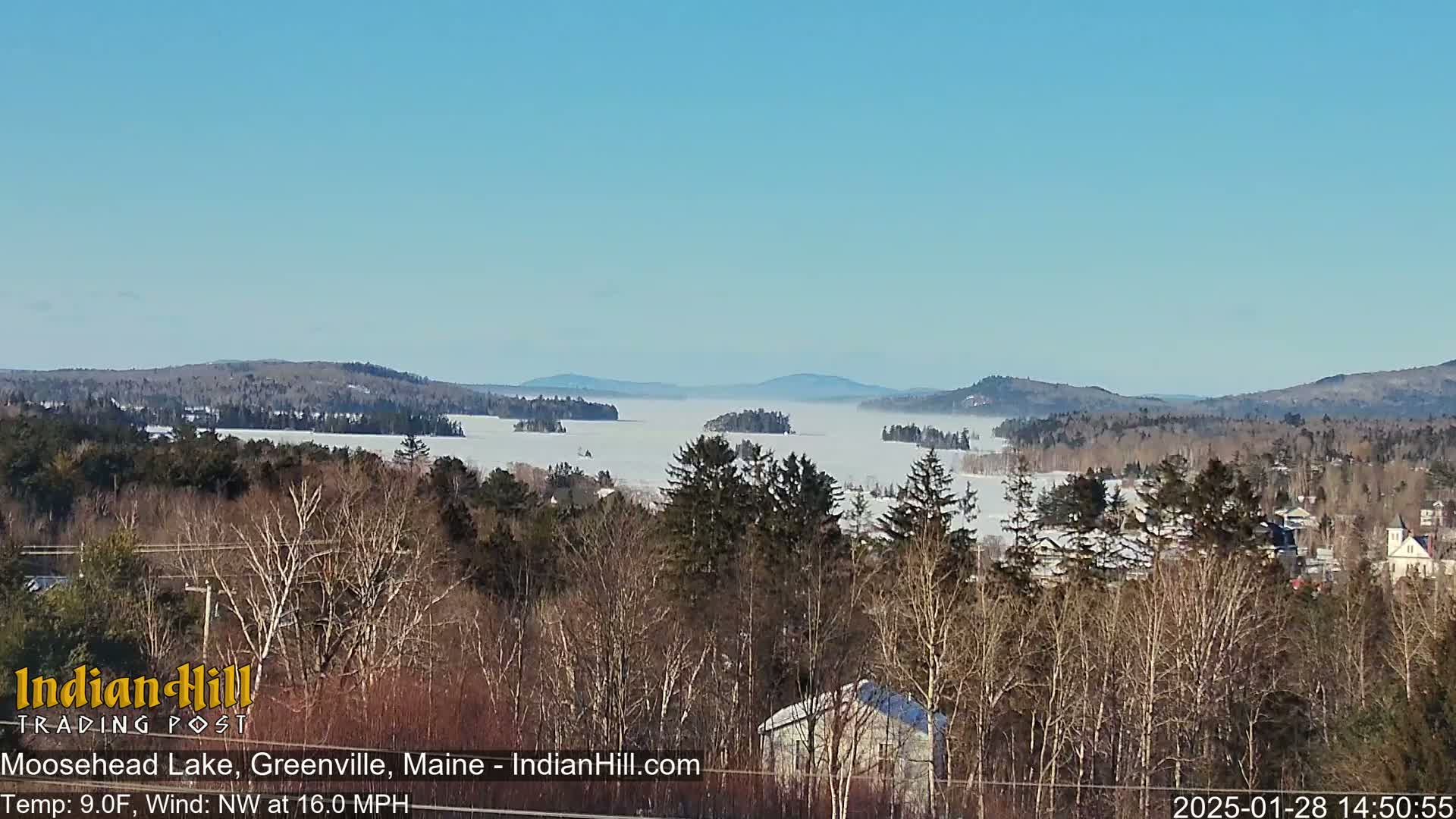 A snow-covered lake stretches to distant, snow-dusted mountains under a clear blue sky, with a layer of fog or mist partially obscuring the water.