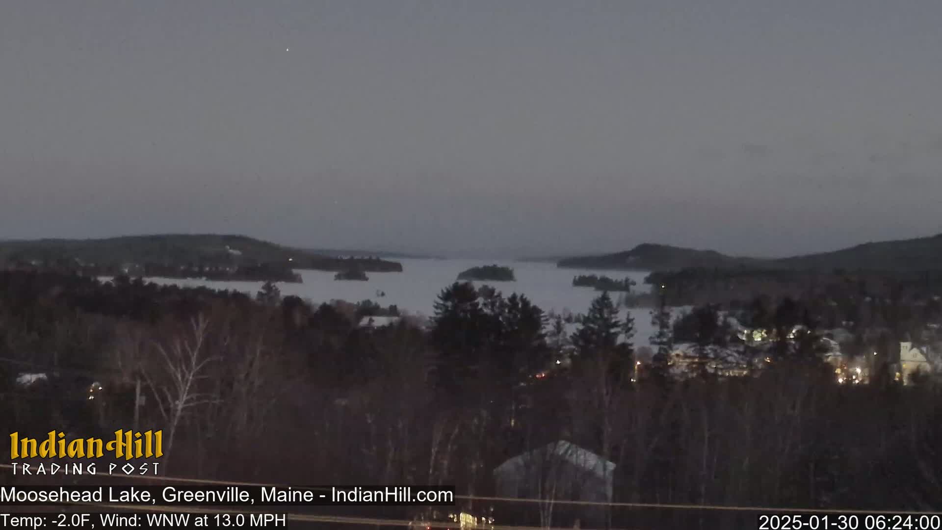 A twilight view of a snow-covered lake with islands, surrounded by dark trees and a small town, under a gray sky.