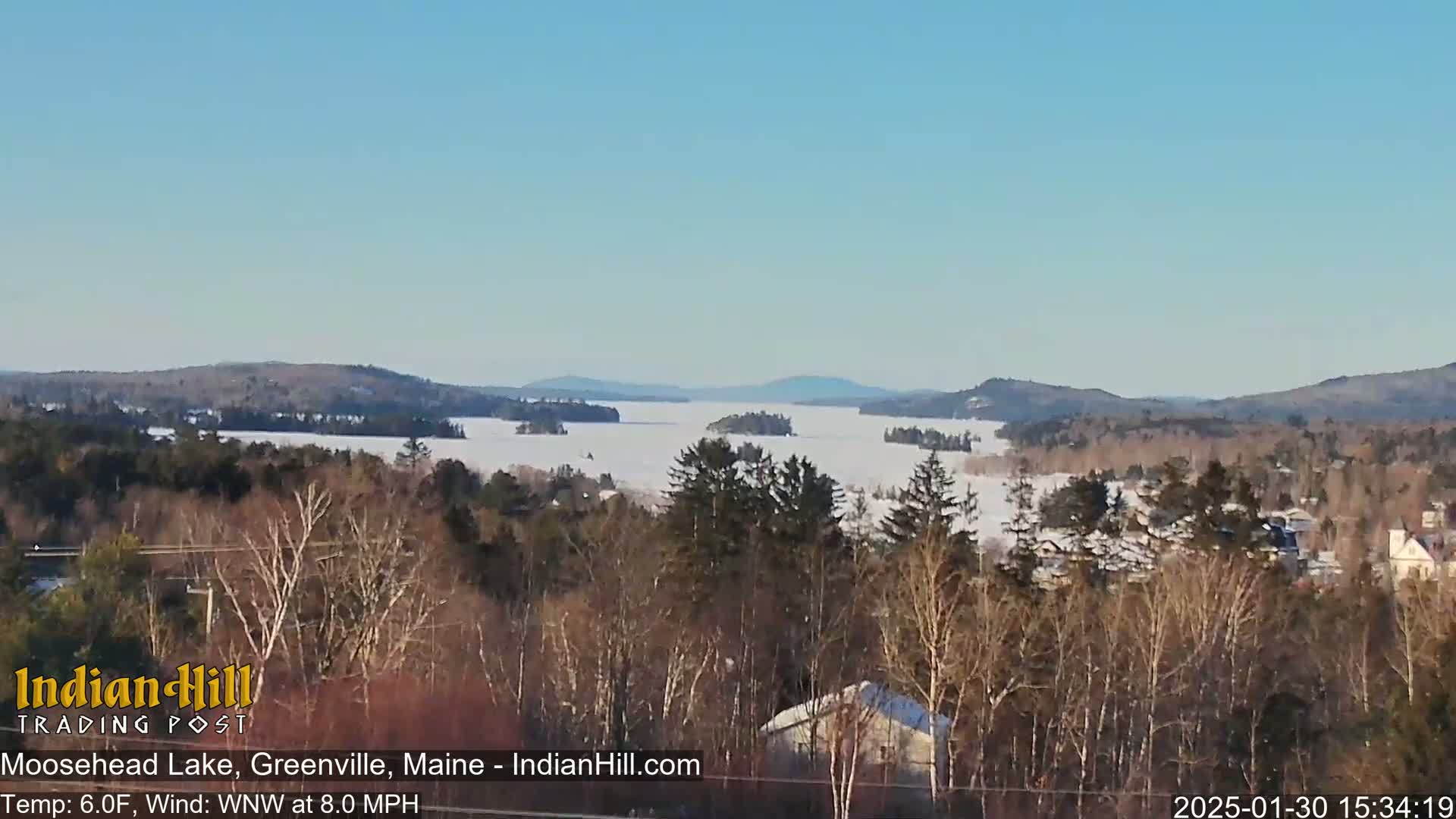 A snow-covered lake stretches to distant hills under a clear blue sky, viewed from a vantage point overlooking a winter landscape of bare trees and houses.