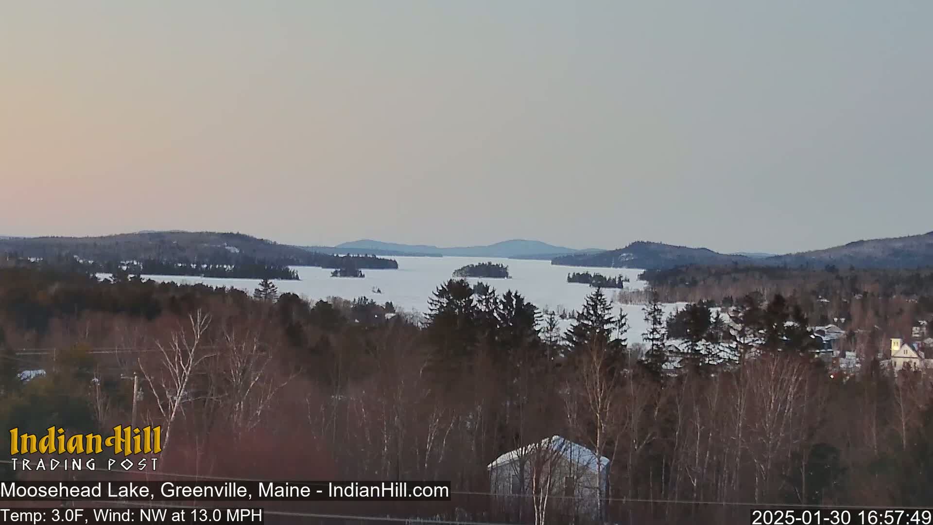 A snow-covered lake with numerous islands is seen from a forested hilltop under a pale, dusky sky.