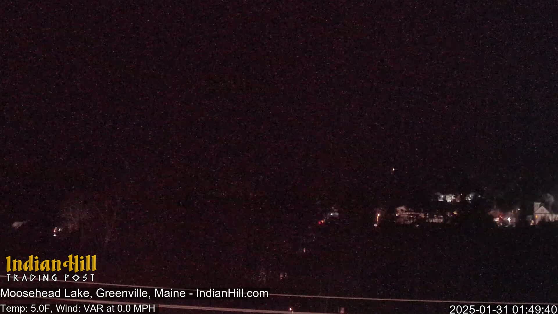 A nighttime long shot of a dimly lit lakeside town under a dark, star-filled sky.